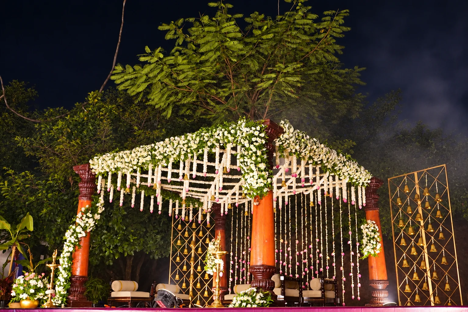 Orange wooden mandap pillars with hanging jasmine and pink lotus flowers at night