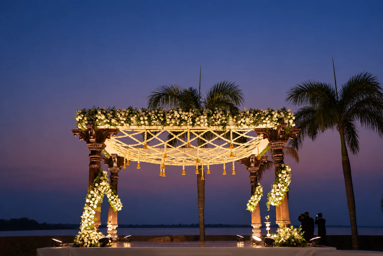 Warmly lit wooden mandap with jasmine canopy beneath a dusk sky
